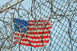 A barbed wire prison fence partially obscuring the USA flag and blue sky in the background 