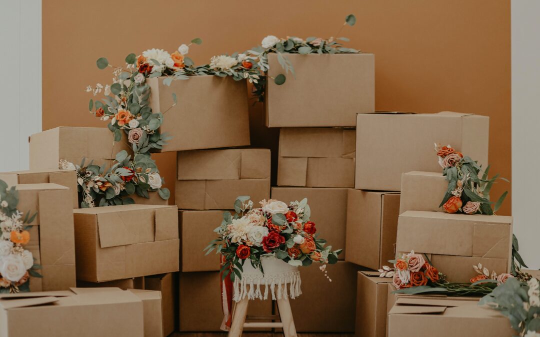 In a room with orange wall, a bunch of boxes are stacked. Some roses with leaves are piled on top of the boxes and a stool with a roses on top is in the bottom middle of the picture.