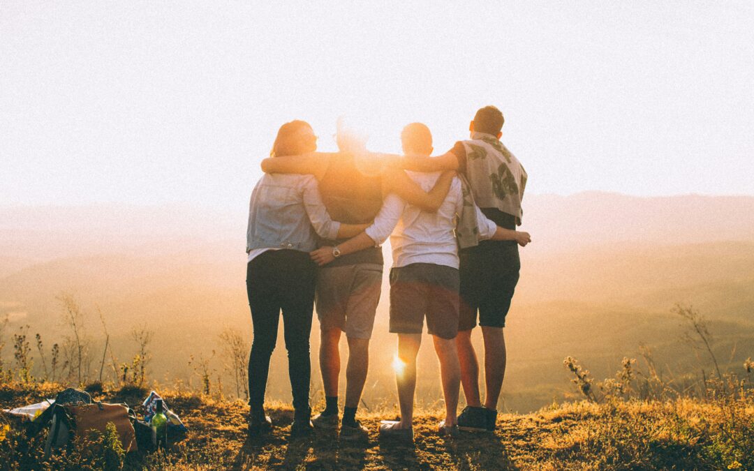 four friends are hugging in a line facing a sunset while standing on a cliff, looks like they finished a hike.