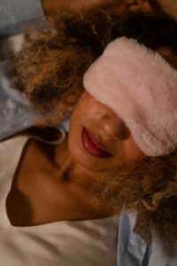 Person sleeps with pink fuzzy face mask. They have brown skin and light brown hair. They wear a white top and their lips are red.