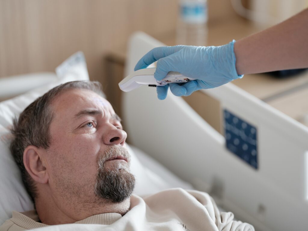 White man in medical setting laying on bed. Someone wearing blue disposable gloves takes his temperature.