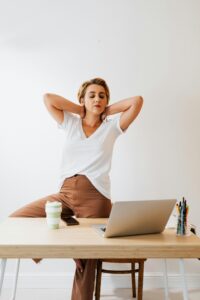 Person with their hands behind their head stretching. They wear a white shirt and brown pants. They sit in front of a desk with a laptop and pens. They are white and have brown and blonde hair.